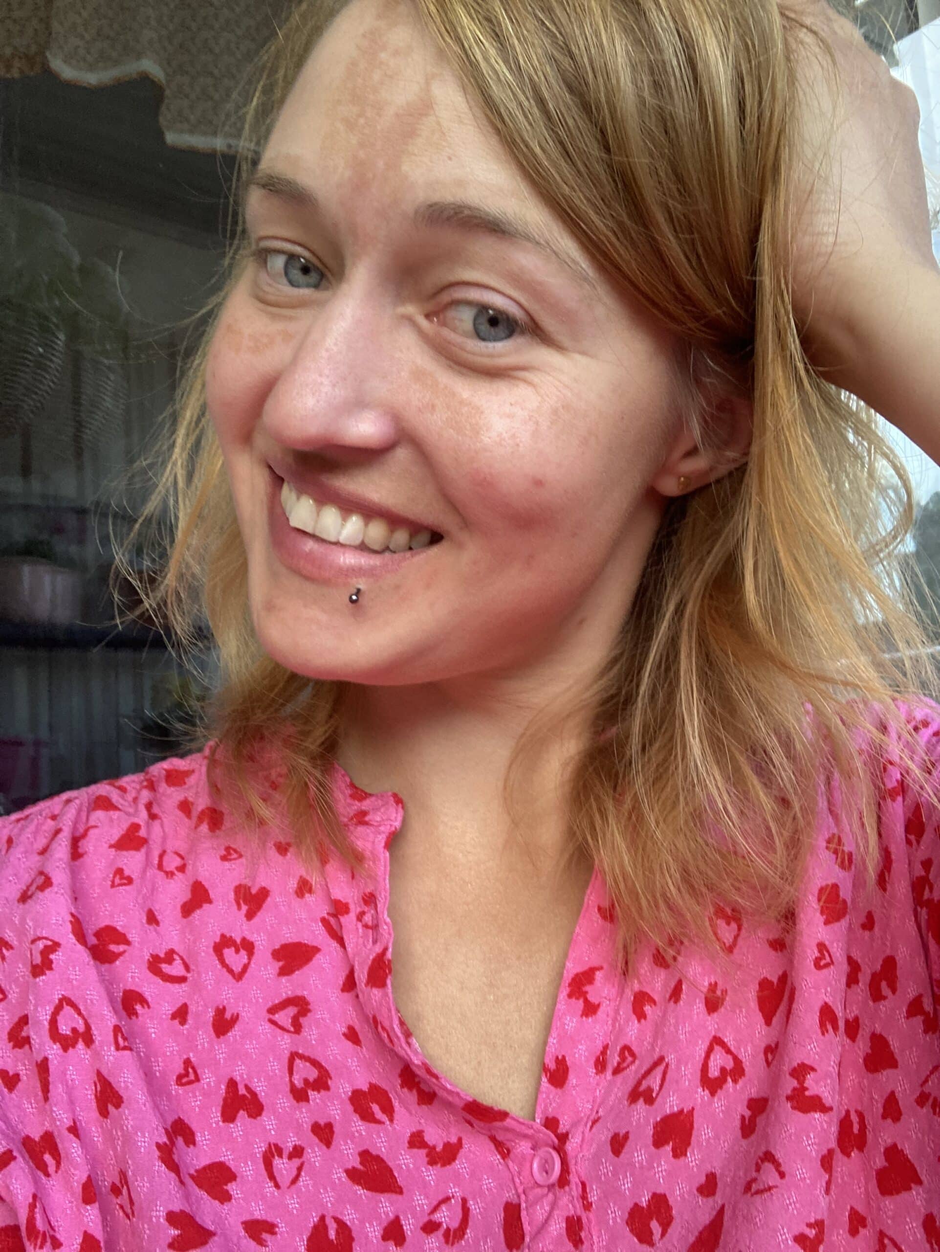 Kvinna i rosa skjorta med röda hjärtan, lutar huvudet mot handen och ler mot kameran, framför en bakgrund med färgglada blommor. Bilden illustrerar en presentation av Martina som coach, med fokus på att hjälpa andra att ta sig igenom separationer med mer mod, självrespekt och klarhet.