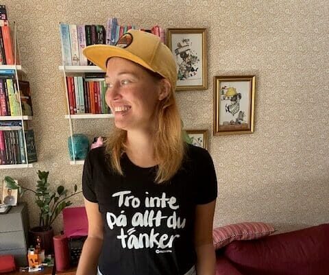 Kvinna i t-shirt med texten “Tro inte på allt du tänker” och keps, står avslappnat med händerna i byxfickorna i ett vardagsrum med bokhyllor bakom sig. Bilden hör till en text om personlig utveckling, inre trygghet, gränssättning och att känna mer energi och närvaro i vardagen.