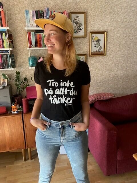 Kvinna i t-shirt med texten “Tro inte på allt du tänker” och keps, står avslappnat med händerna i byxfickorna i ett vardagsrum med bokhyllor bakom sig. Bilden hör till en text om personlig utveckling, inre trygghet, gränssättning och att känna mer energi och närvaro i vardagen.